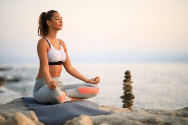 Young woman with eyes closed doing breathing exercises while meditating on a rock at the beach. Copy space. 