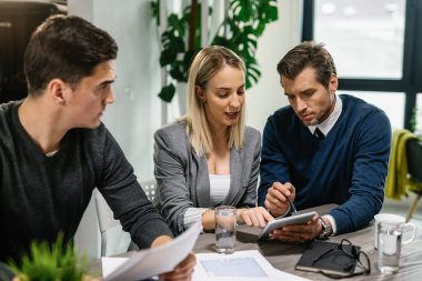Rear estate agent and young couple going through housing plans on touchpad while having a meeting at home. 