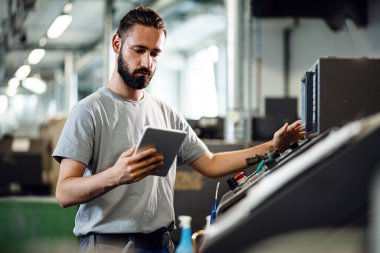 Young engineer using touchpad while working at CNC machine in a factory.