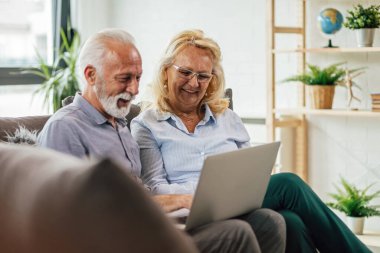 Happy senior couple using laptop and watching something on the internet while sitting on the sofa in the living room.