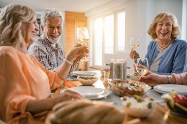 Happy seniors friends having lunch together and toasting with wineglasses at dining table at home. 