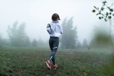 Back view of female runner jogging in nature on foggy morning. Copy space.