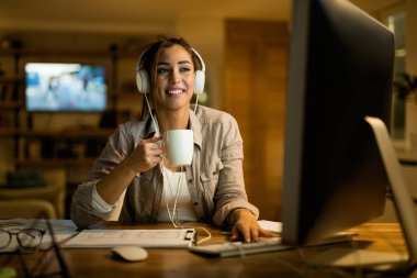 Young smiling woman wearing headphones while using computer and drinking coffee in the evening at home. 