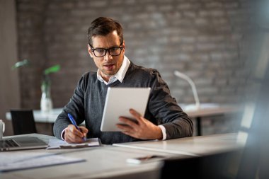 Male entrepreneur writing business report while using digital tablet in the office. 