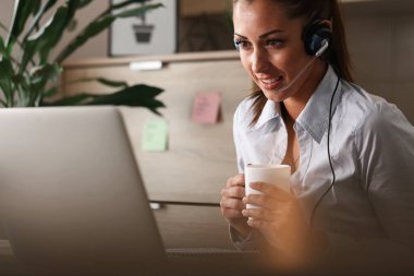 Smiling customer service representative with headset using a computer while talking to a customer and drinking coffee in call center. 