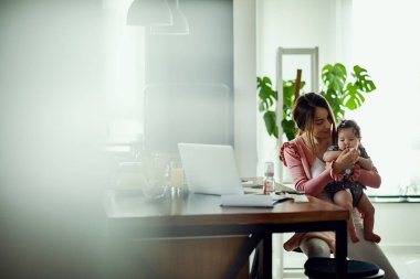 Happy mother giving a pacifier to her baby daughter while working on a computer at home. 