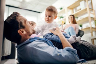 Caring father lying on the floor and holding his baby boy. Mother and daughter are in the background. 