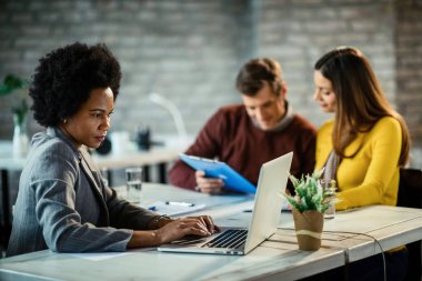Black female insurance agent using computer while working on a meeting in the office. Her clients are in the background. 