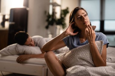 Young woman with smart phone contemplating and feeling worried about her relationship while boyfriend is sleeping on the bed. 