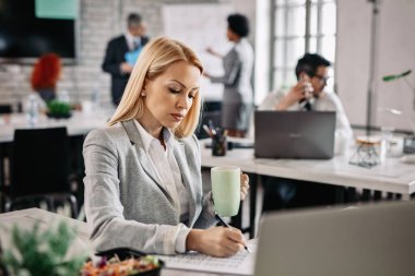 Concentrated female entrepreneur writing notes while analyzing business reports in the office. There are people in the background. 