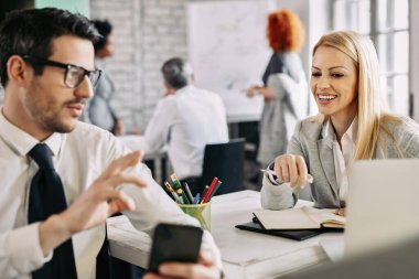 Happy mid adult businesswoman having fun while her colleague is showing something on smart phone to her in the office. 