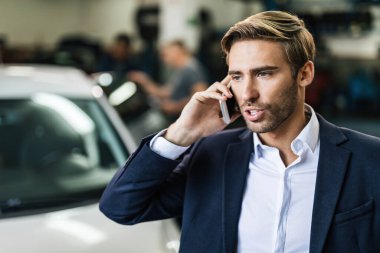 Young businessman communicating on mobile phone while auto mechanics are repairing his car in the background at workshop. 
