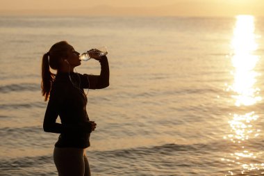 Thirsty athletic woman drinking water from a bottle and refreshing herself by the sea at dawn. Copy space. 