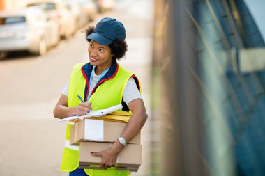 Smiling African American courier making a delivery and filling documents while standing next to her mini van. 