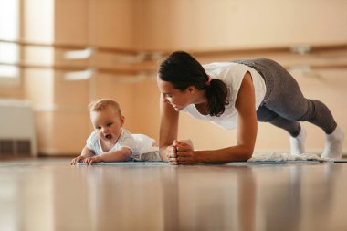 Young sportswoman in plank position exercising with her baby boy in health club. Focus is on baby. Copy space. 