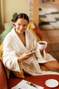 Young smiling woman in bathrobe drinking tea and using smart phone while relaxing at wellness center. 