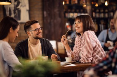 Group of happy friends communicating while relaxing in coffee shop. Focus is on happy woman holding her eyeglasses. 