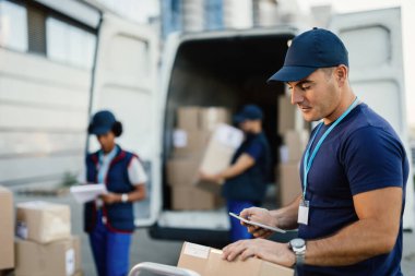 Young delivery man checking package while using digital tablet outdoors. His coworkers are unloading boxes from a van in the background. 