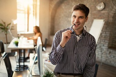 Portrait of young smiling businessman standing in the office. His colleague is working in the background. 