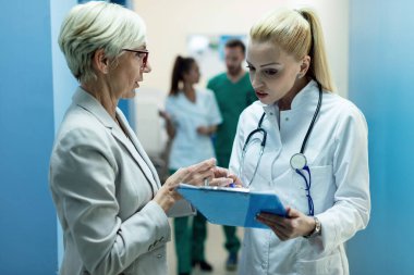 General practitioner and mature woman analyzing medical records and talking about test results in a hallway at clinic. 