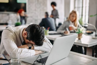 Tired entrepreneur sleeping on the table after working on computer in the office. His colleagues are working in the background. 