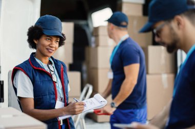 Happy African American worker communicating with her colleague while writing on clipboard during package delivery. 