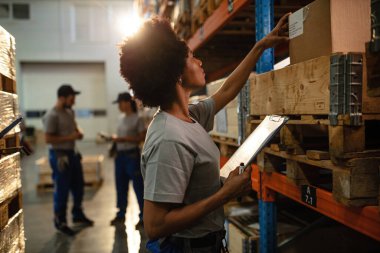 Black female warehouse worker holding shipment list while checking packages on the shelf in industrial storage compartment. 