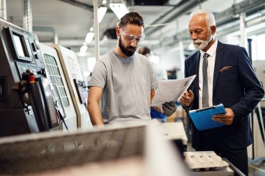Factory worker and mature engineer examining product report in industrial facility. 