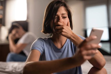 Sad woman feeling disappointed while reading text messages on unfaithful boyfriend's mobile phone at home. The man is in the background. 