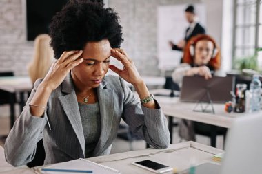 African American businesswoman looking stressed while holding her head in pain in the office. There are people in the background. 