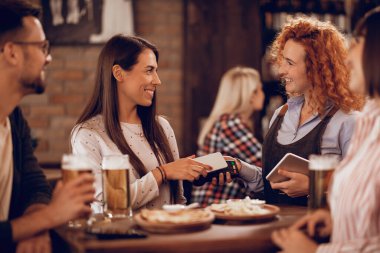 Happy waitress holding electronic reader while her customer is paying with smart phone via contactless payment. 