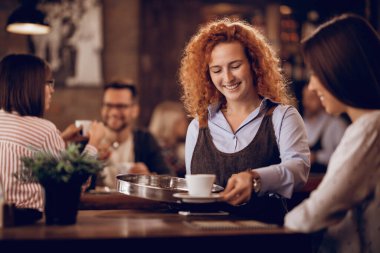 Young happy waitress giving coffee to a female guest while working in a bar. 