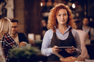 Portrait of female student with digital tablet working part-time as a waitress in a pub. 