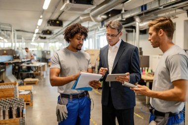 Mid adult engineer and metal workers cooperating while analzying production reports in industrial building. 