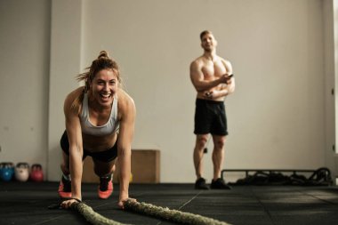 Cheerful female athlete doing push-ups while exercising with battle rope and having fun in a gym There is a man in the background. 