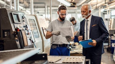 Manual worker and senior businessman cooperating while going through paperwork in factory plant. 