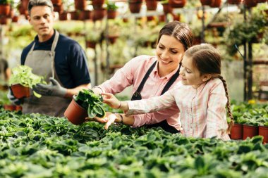 Happy mother working with her daughter at plant nursery and teaching her how to take care on flowers. 