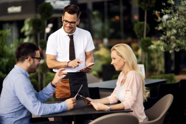 Young happy waiter communicating with guests while taking their order on a touchpad in a cafe. 