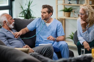 General practitioner examining senior man with a stethoscope and listening to his heartbeat during a home visit. 