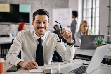 Young happy businessman working in the office and looking at camera. There are people in the background. 