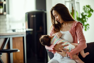 Smiling mother breastfeeding her baby daughter while being at home. 