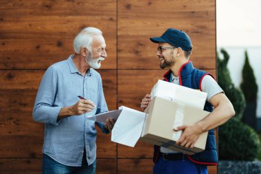 Happy delivery man talking with mature customer who is signing delivery documents. 