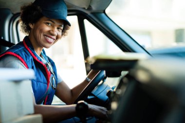 Happy black female courier driving delivery van and looking at camera. 