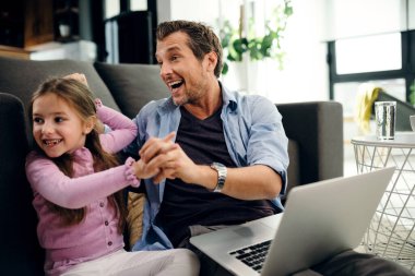 Happy father and daughter having fun while holding hands and using laptop in the living room. 