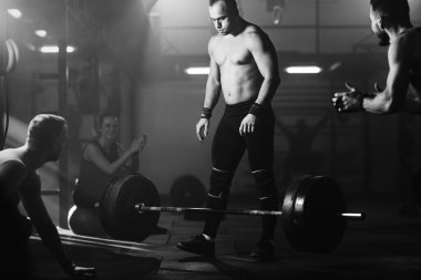 Black and white photo of shirtless sportsman concentrating for deadlift while his friends are supporting him in a gym. 