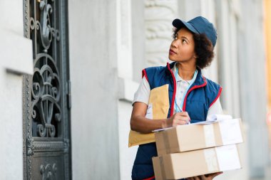 African American female deliverer writing notes while dispatching packages in the city. 