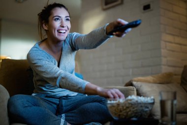 Young happy woman changing channels with remote control while watching TV and eating popcorn in the evening at home. 
