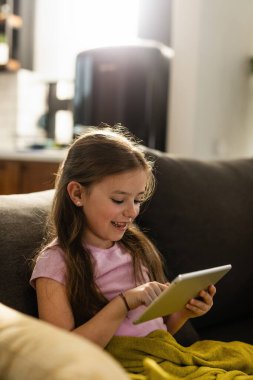 Happy little girl using touchpad and playing games while relaxing on the sofa at home. 