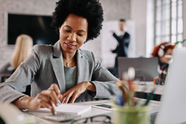 Smiling black entrepreneur working on financial reports at her desk in the office. There are people in the background. 