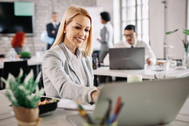 Happy female entrepreneur using laptop while working at her office desk. There are people in the background. 